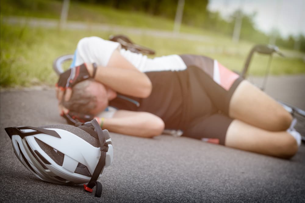 Injured cyclist on the pavement clutching his head, with a helmet in the foreground.