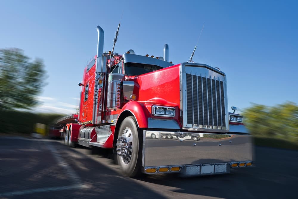 Red classic American big rig semi truck with stylish chrome accessories parked at a truck stop, carrying covered commercial cargo on a flatbed trailer.