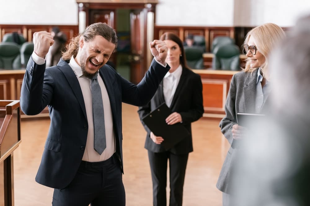 Energetic man celebrates victory, shouting with a triumphant gesture beside his attorney in a courtroom.