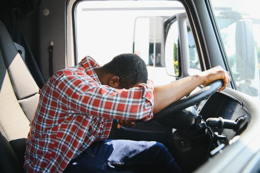 Exhausted from work, a young African American truck driver rests inside his cabin, parked alongside his vehicle during daylight hours.