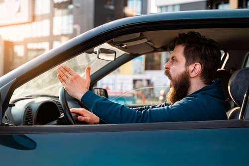 Bearded man expressing frustration and road rage while driving in his car, gesturing with his hand.
